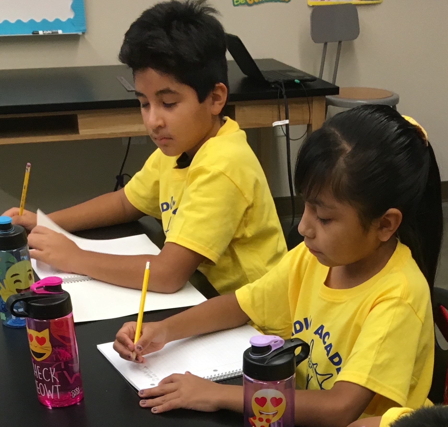 Two students in yellow Reading Academy shirts writing at a classroom desk, 2017