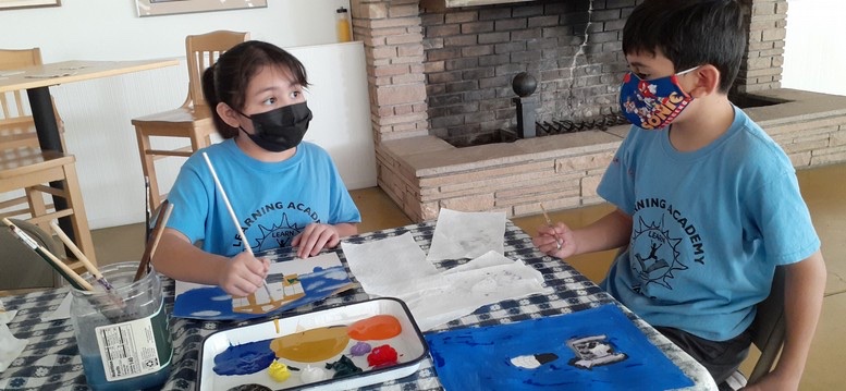 Two masked Learning Academy students painting at a classroom table, 2021