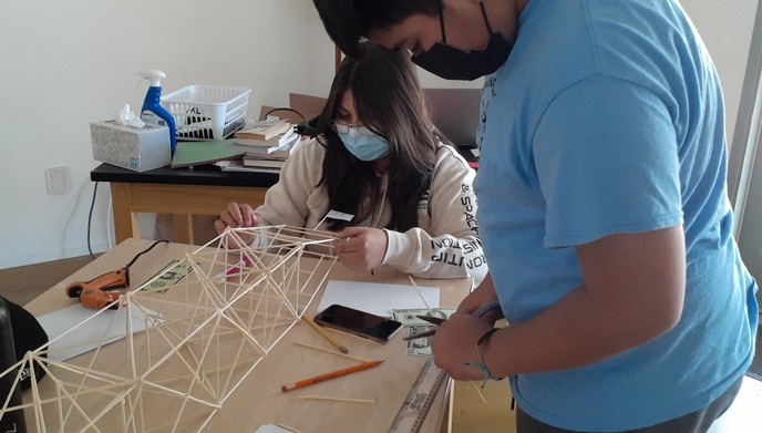 Two masked Learning Academy students working on a toothpick bridge-building project