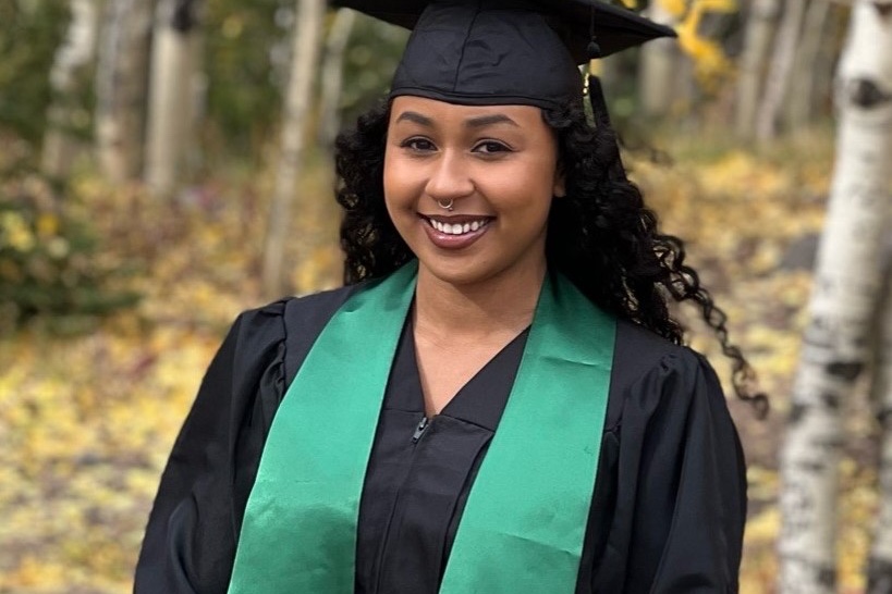 A BASIC scholar in cap and gown with her Cal Poly Humboldt stole, among autumn aspens