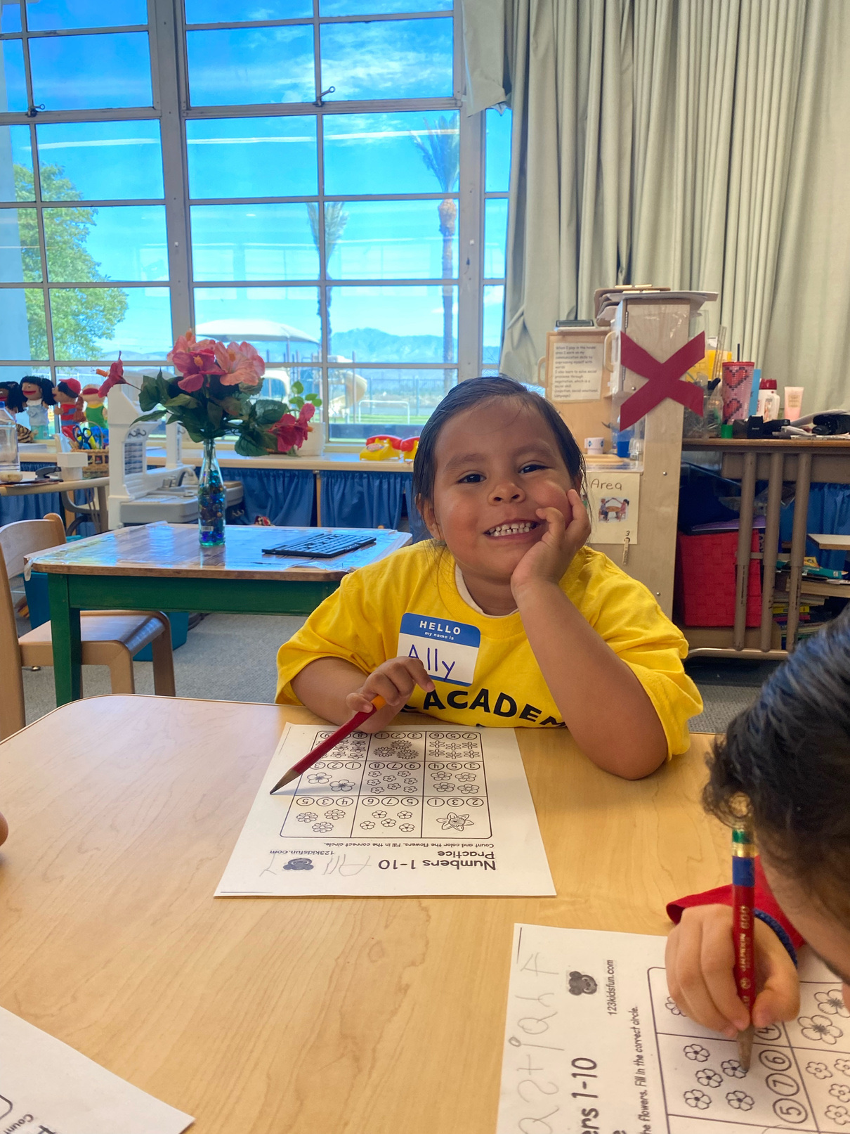 A TK Academy student in a yellow program t-shirt smiling at her desk over a numbers worksheet