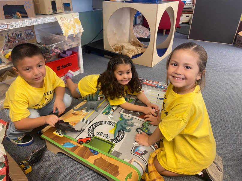 Three TK Academy students playing together with toy dinosaurs on a play mat