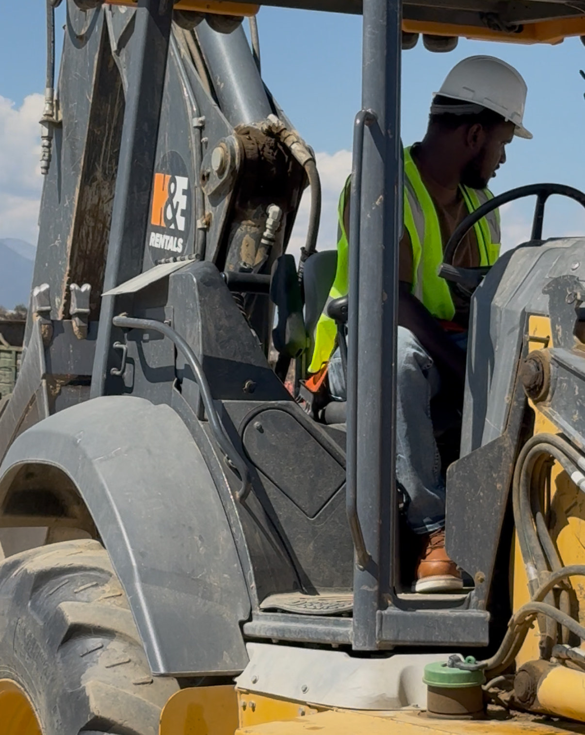 Briem Cole in a hard hat and high-visibility vest at the controls of a backhoe loader on a work site