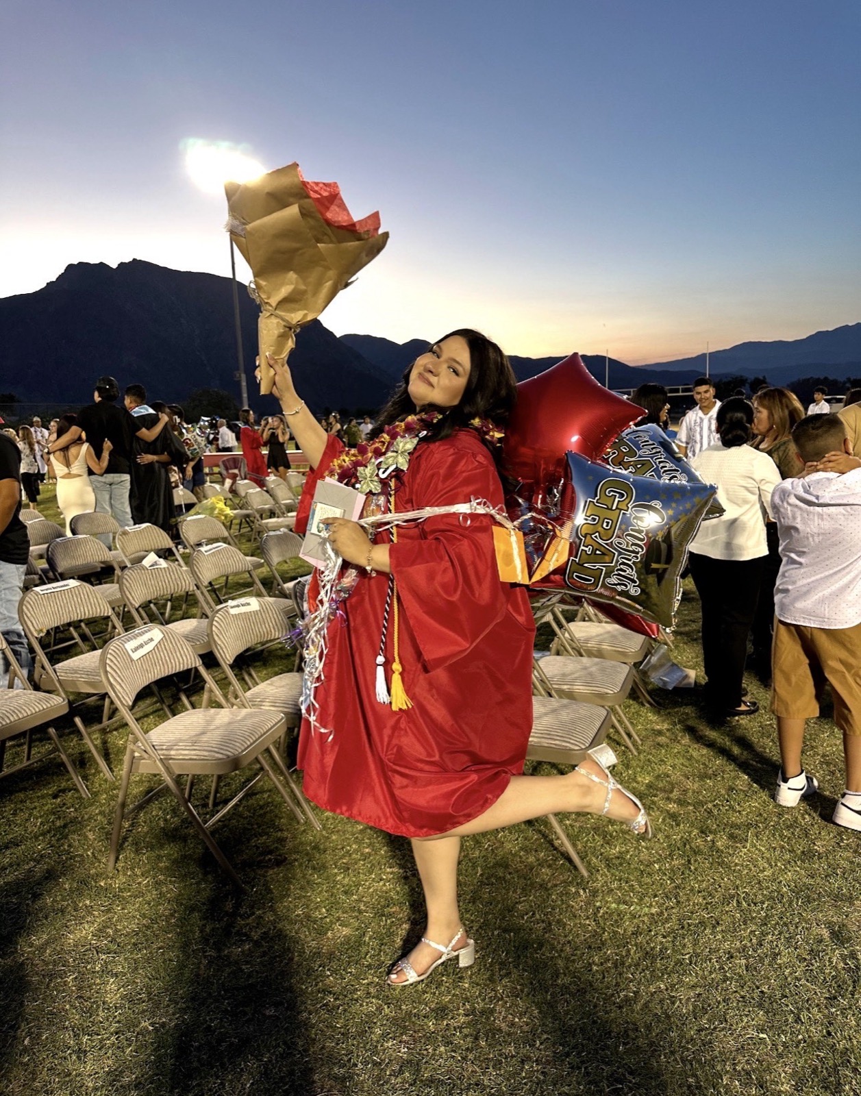 Jimena Ochoa in cap and gown holding a bouquet and balloons above her head at an outdoor graduation, mountains behind