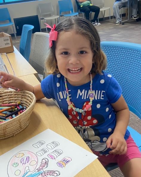 A young girl at a table coloring a picture, smiling at the camera