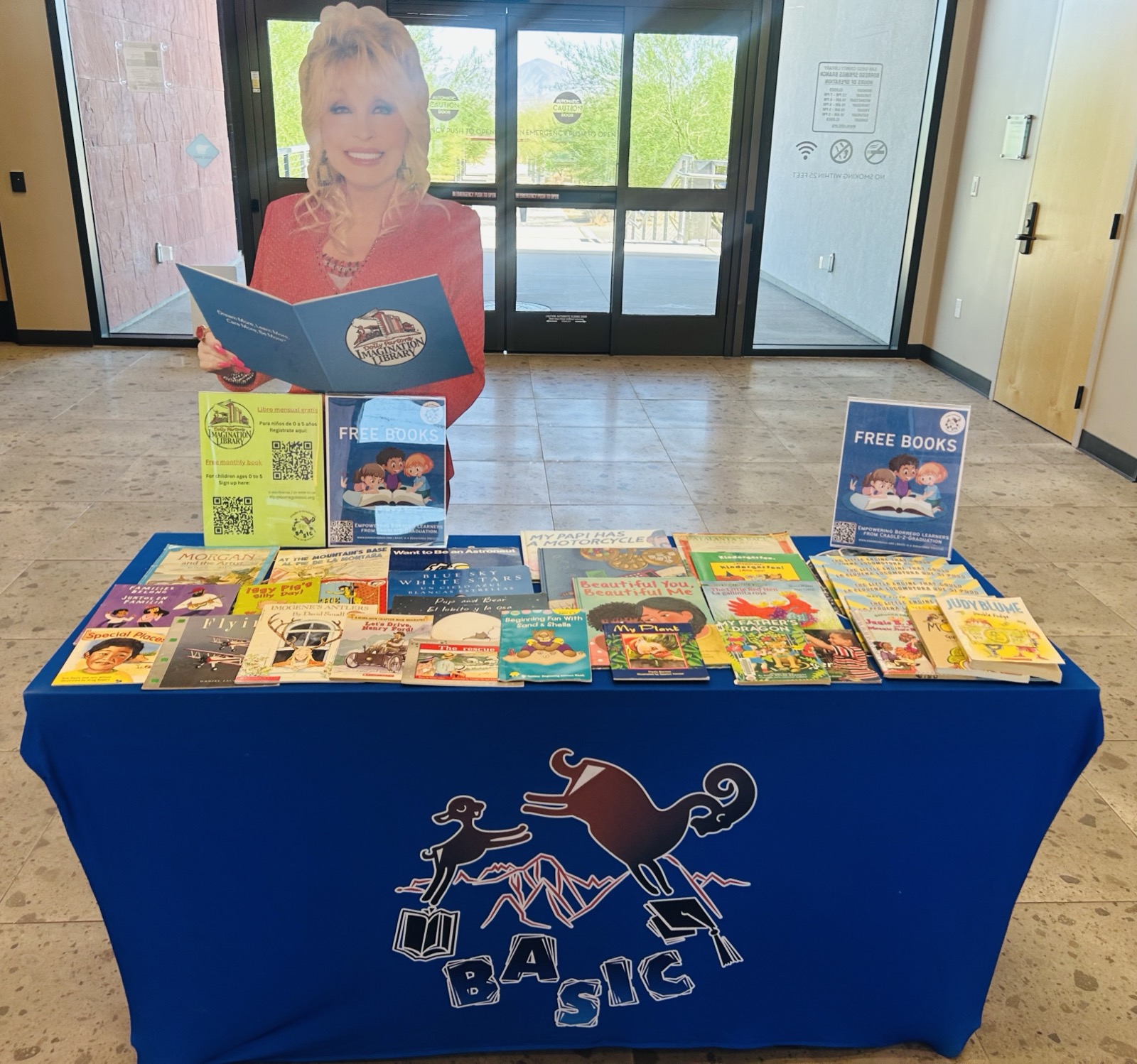 BASIC's Free Book Table in the lobby of the Borrego Springs Library: two long rows of children's picture books in English and Spanish on a blue BASIC tablecloth, with bilingual Imagination Library sign-up flyers and a Dolly Parton cutout behind