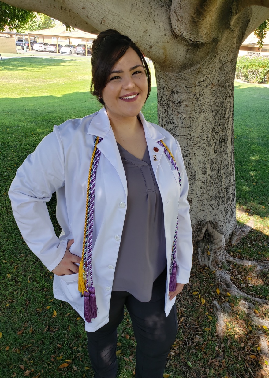Jamie Blakeley in a white clinical coat and purple honor cords beneath a tree