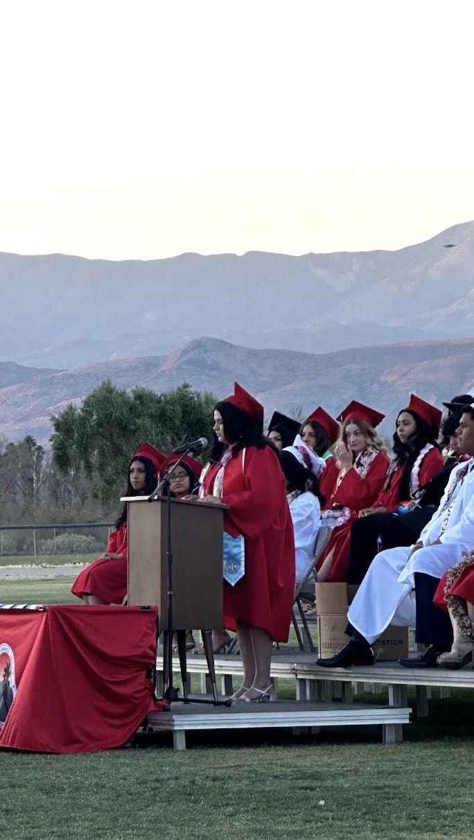 Jimena Ochoa speaking at the podium during the Borrego Springs High School graduation, classmates in red seated behind her
