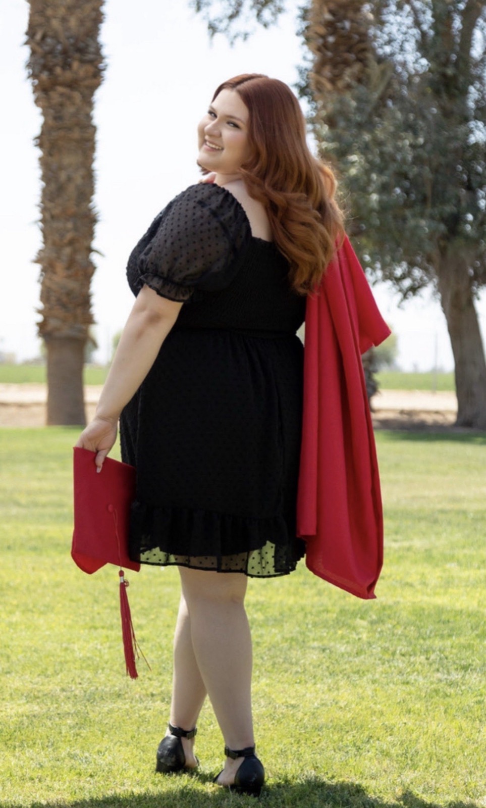 Jimena Ochoa in a red graduation gown, cap in hand, on a lawn lined with palm trees