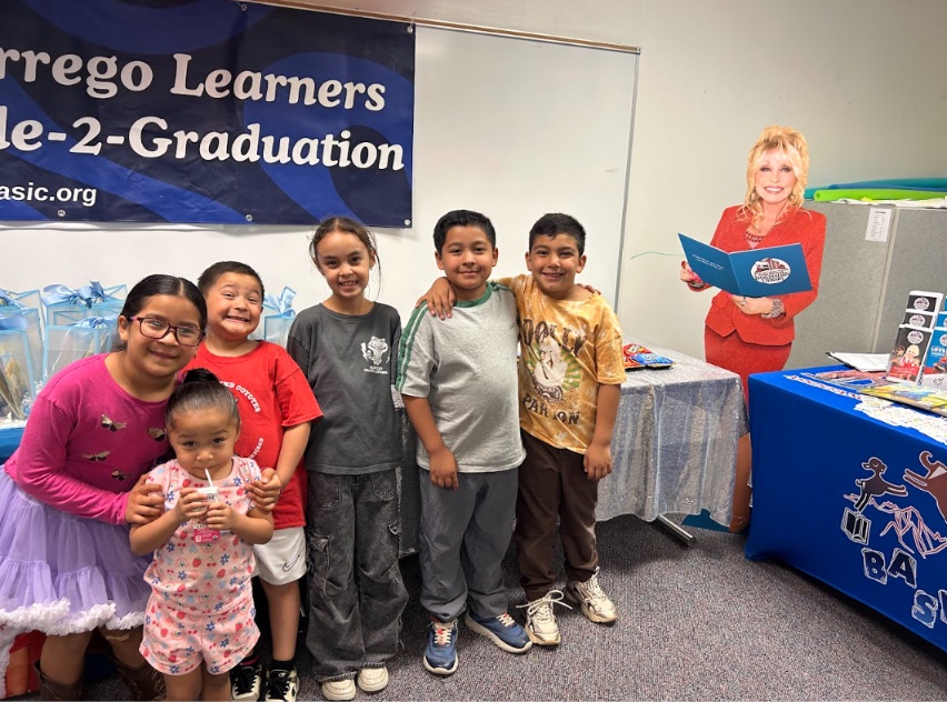 Six Borrego Springs children standing together and smiling in front of a BASIC banner reading Empowering Borrego Learners from Cradle-2-Graduation, with a life-size Dolly Parton cutout holding an open book behind them