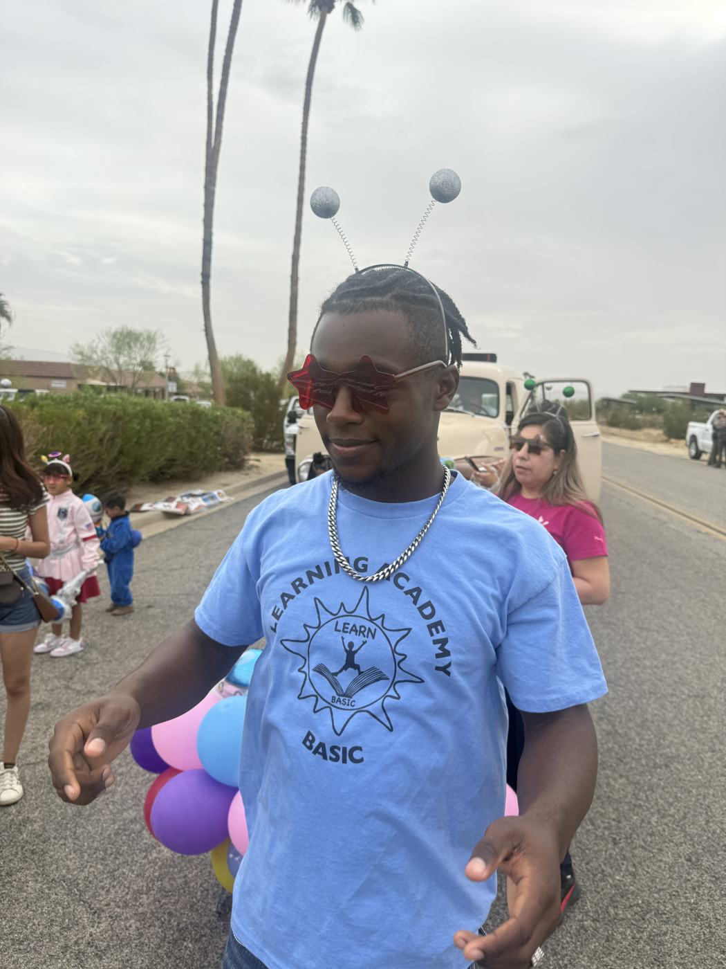 A Learning Academy volunteer in a blue BASIC shirt wearing a silver antenna headband and red star-shaped sunglasses, with a bunch of balloons behind him