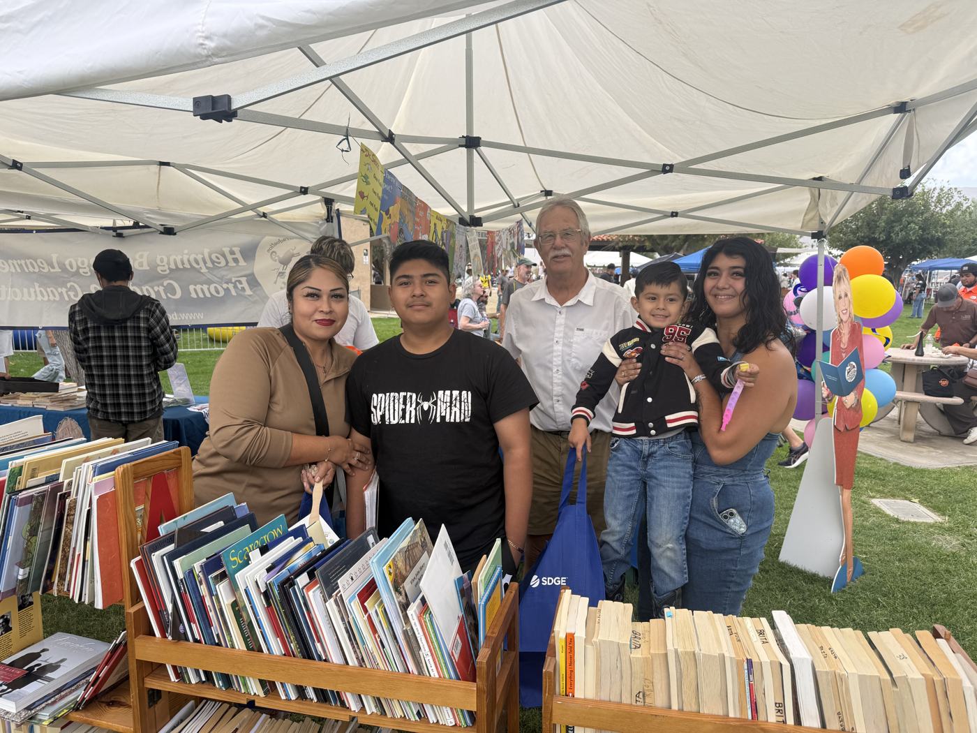 Dick Fausel at the free book table under the BASIC canopy with a family of four holding their chosen books and a blue SDG&E bag