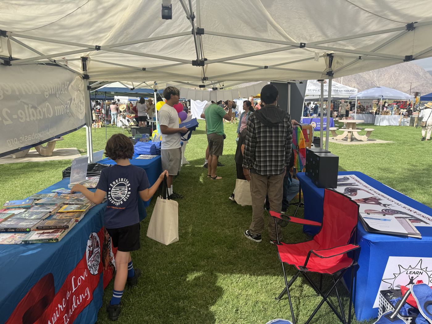 A boy in a navy shirt looking through the rows of free children's books on the blue BASIC table, with festival-goers under the canopy behind him