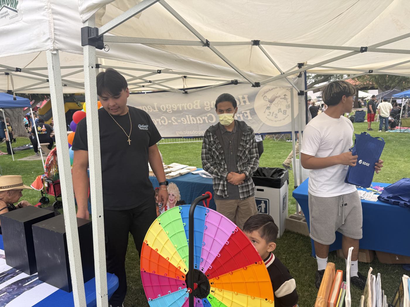 Two Learning Academy volunteers beside the rainbow prize wheel at the BASIC booth, with a small child reaching for it