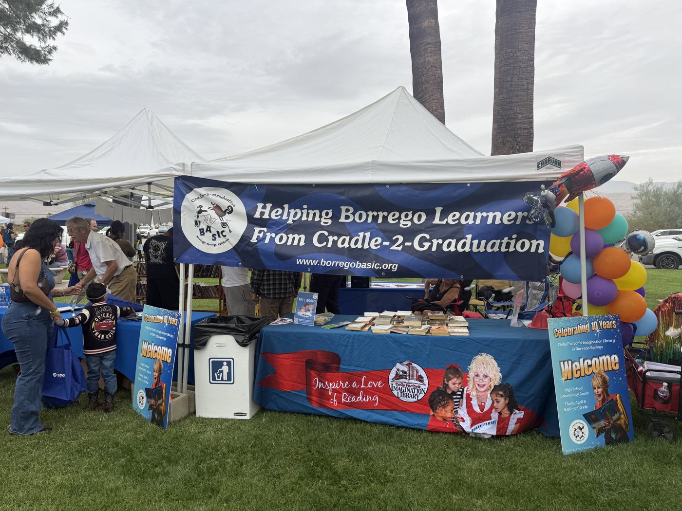 The BASIC booth at Christmas Circle: a white canopy with the banner Helping Borrego Learners From Cradle-2-Graduation, tables of free books, a balloon arch, and Imagination Library signs reading Celebrating 10 Years