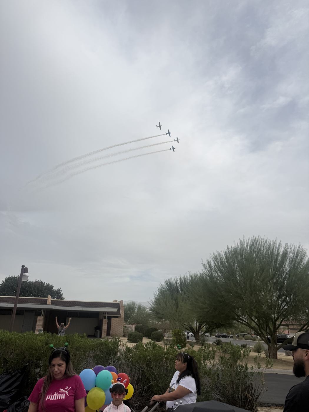 Four small planes trailing smoke over the parade lineup, with desert trees, rooftops and families holding balloons below