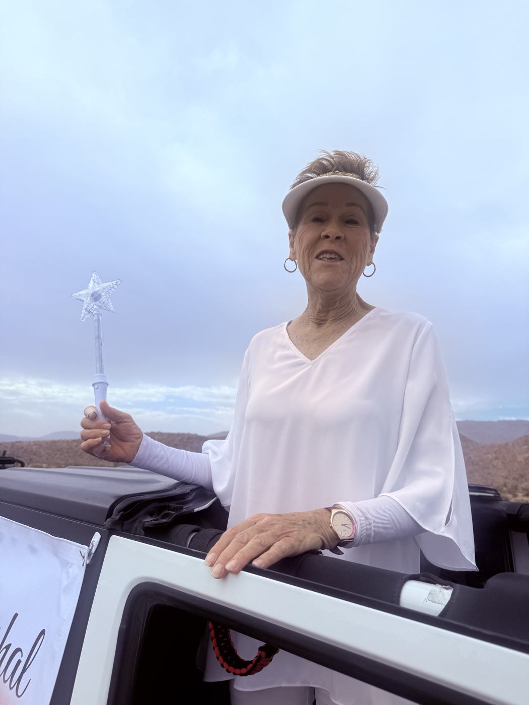 Martha Deichler standing in the back of a white Jeep in a white visor and blouse, holding a lighted star wand, with desert mountains behind her