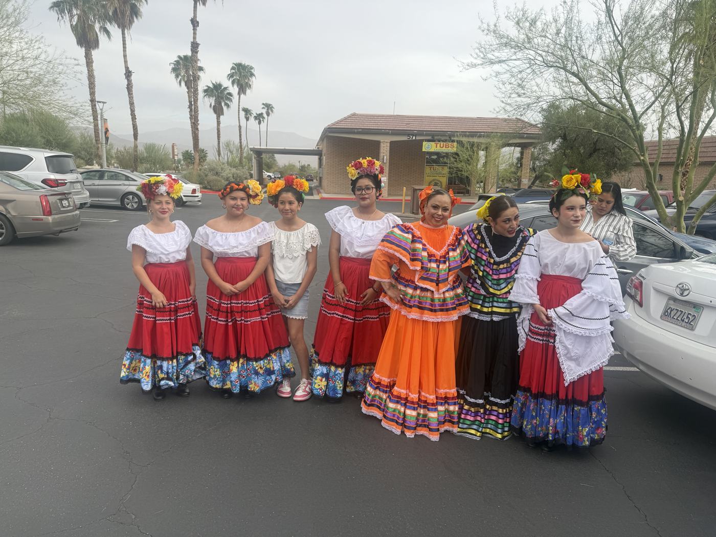Eight folklorico dancers in flowered headpieces and full ruffled skirts in red, orange, blue and purple, lined up in the parking lot before the parade