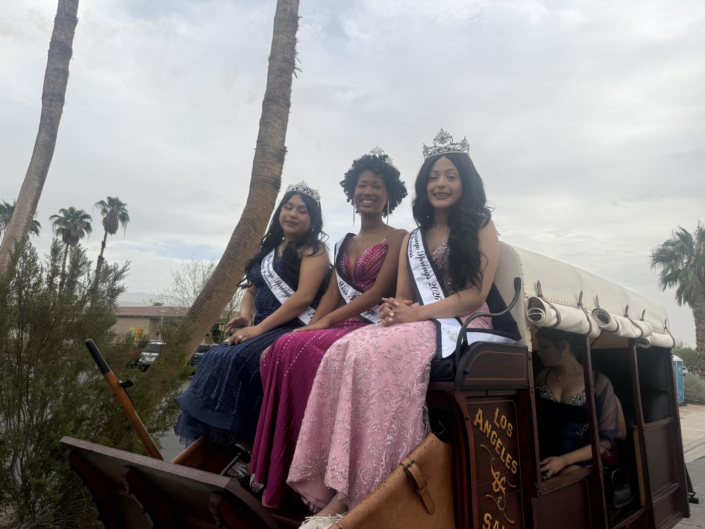 Three members of the Miss Borrego Springs court in crowns, sashes and gowns of navy, magenta and pink, seated on top of a stagecoach