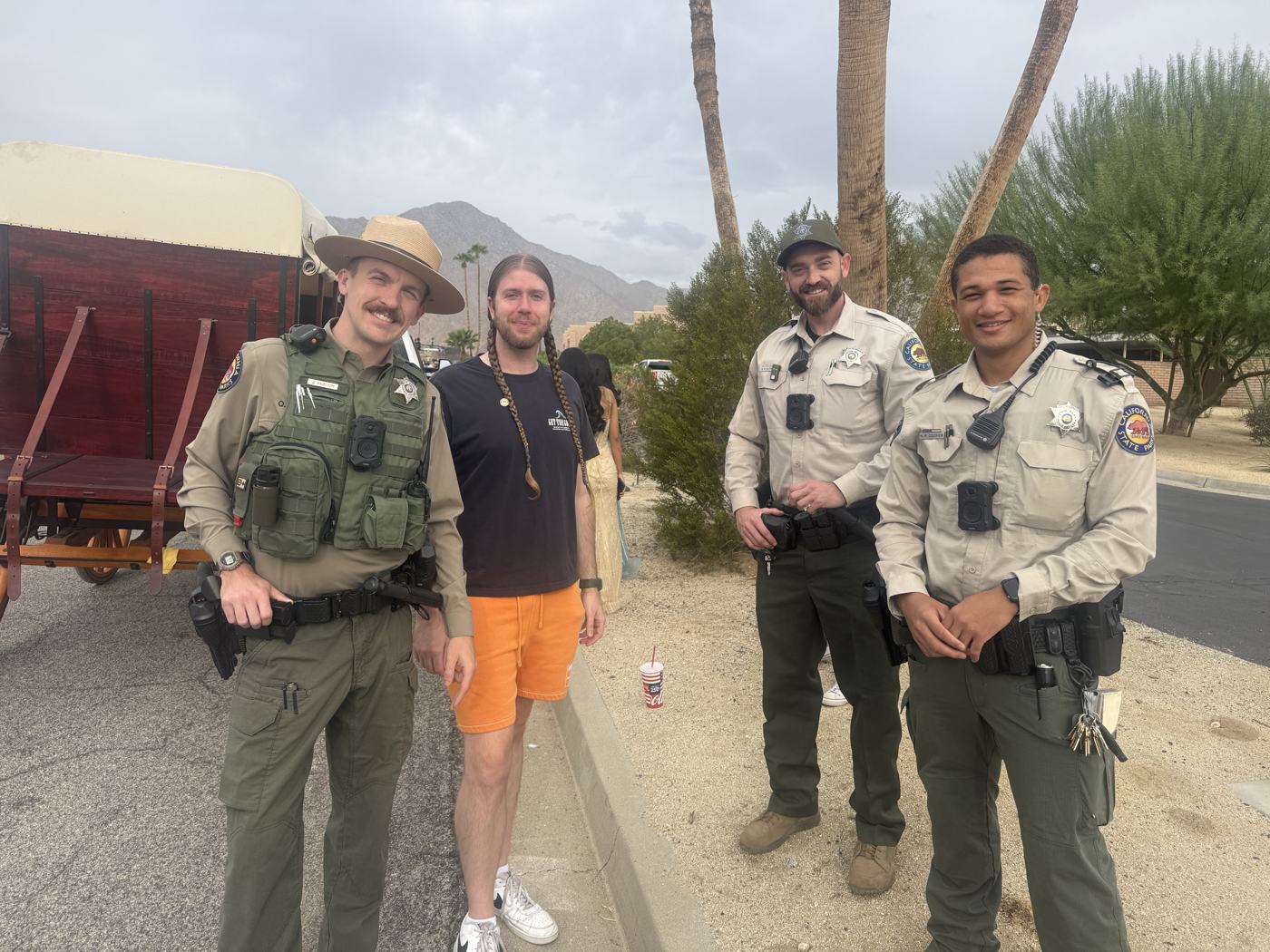 Four uniformed State Parks officers in tan shirts and campaign hats standing with a festival volunteer beside a covered wagon in the parade lineup