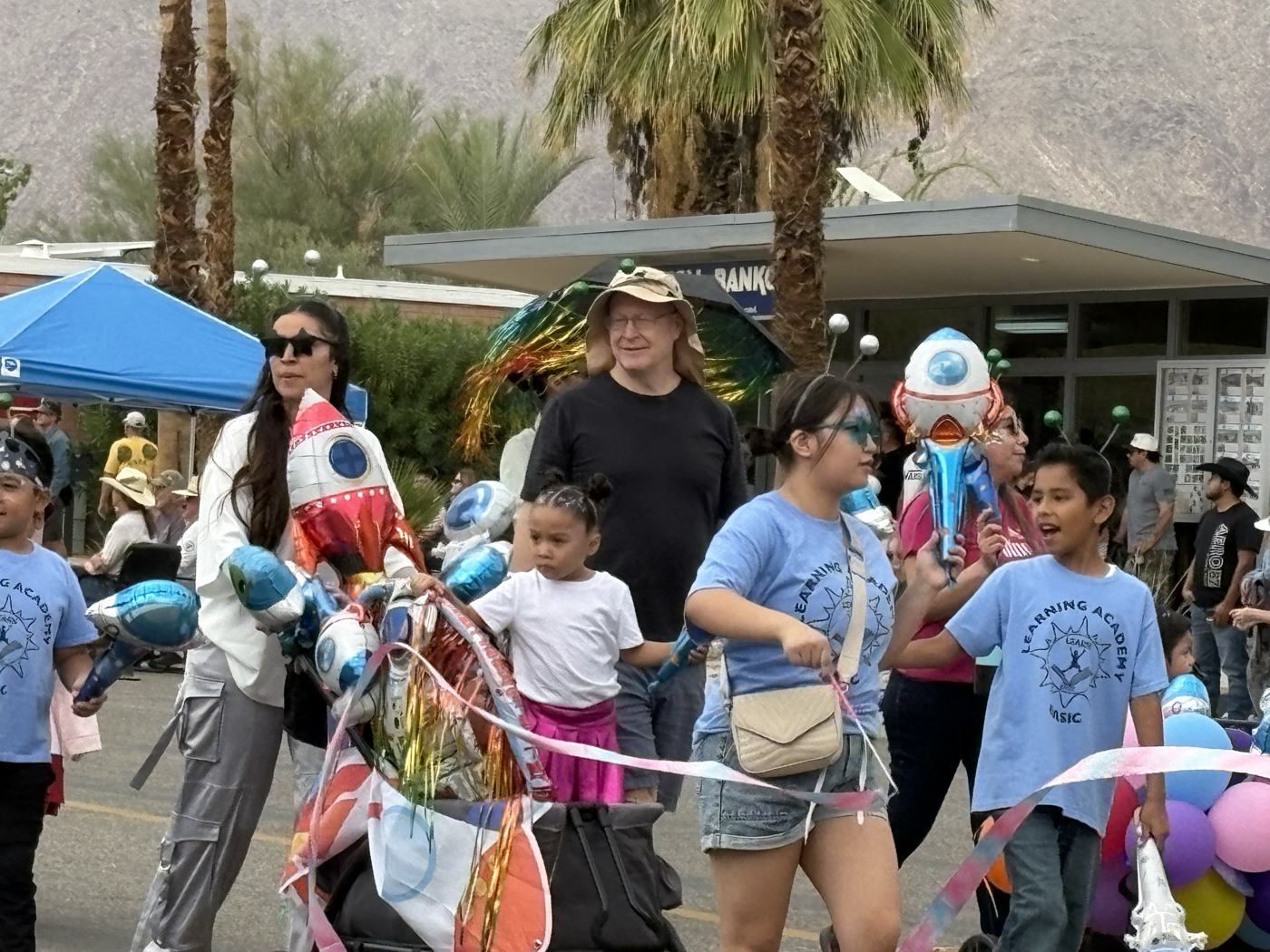 BASIC board member Peter Kellner in a tan hat and antennae walking the parade route alongside children in blue Learning Academy shirts carrying balloons and streamers