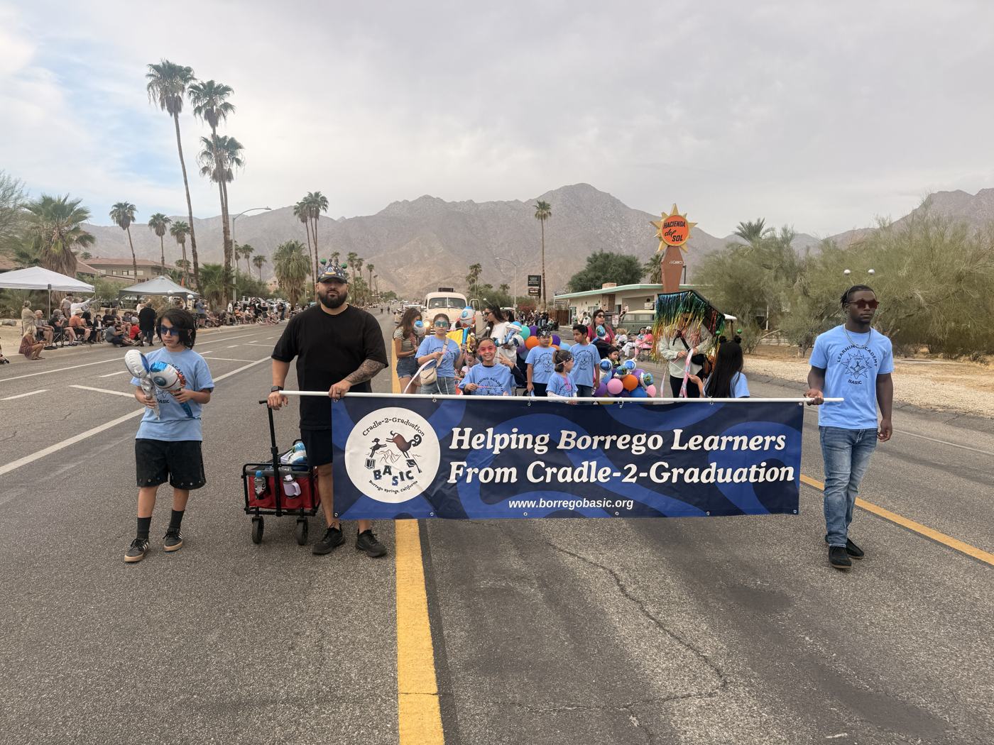 Learning Academy students in light-blue BASIC shirts carrying the banner Helping Borrego Learners From Cradle-2-Graduation down Palm Canyon Drive, with balloons behind them and the Santa Rosa Mountains in the distance