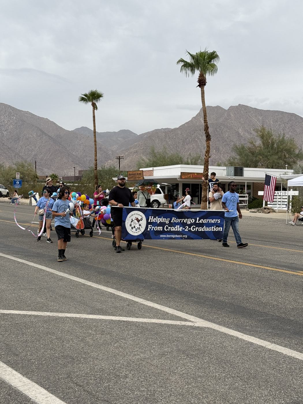The BASIC parade entry seen from the curb: the banner, a wagon, and children with balloons walking past storefronts with a palm tree and mountains behind