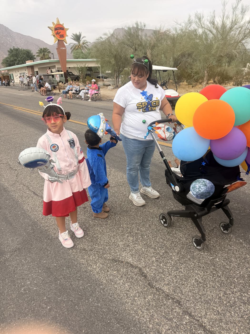 A young girl in a white and red outfit holding a balloon, beside a small boy in a blue jumpsuit and a woman pushing a stroller hung with balloons