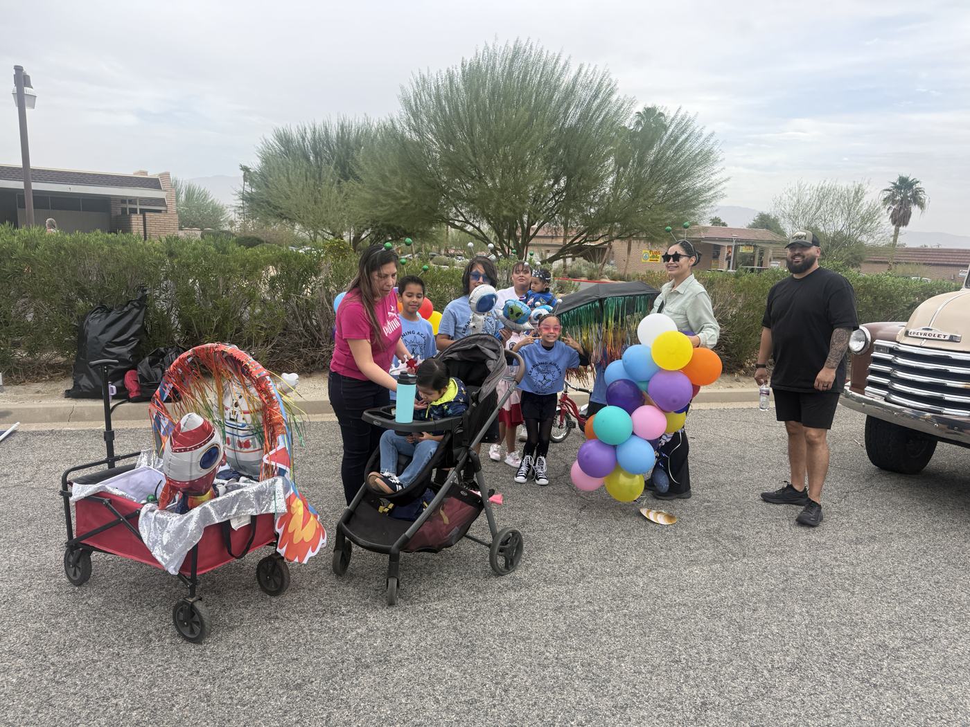 Flor Gomez in star-shaped sunglasses and antennae holding a bunch of balloons, gathering Learning Academy students and parents around the decorated wagon and stroller before the parade