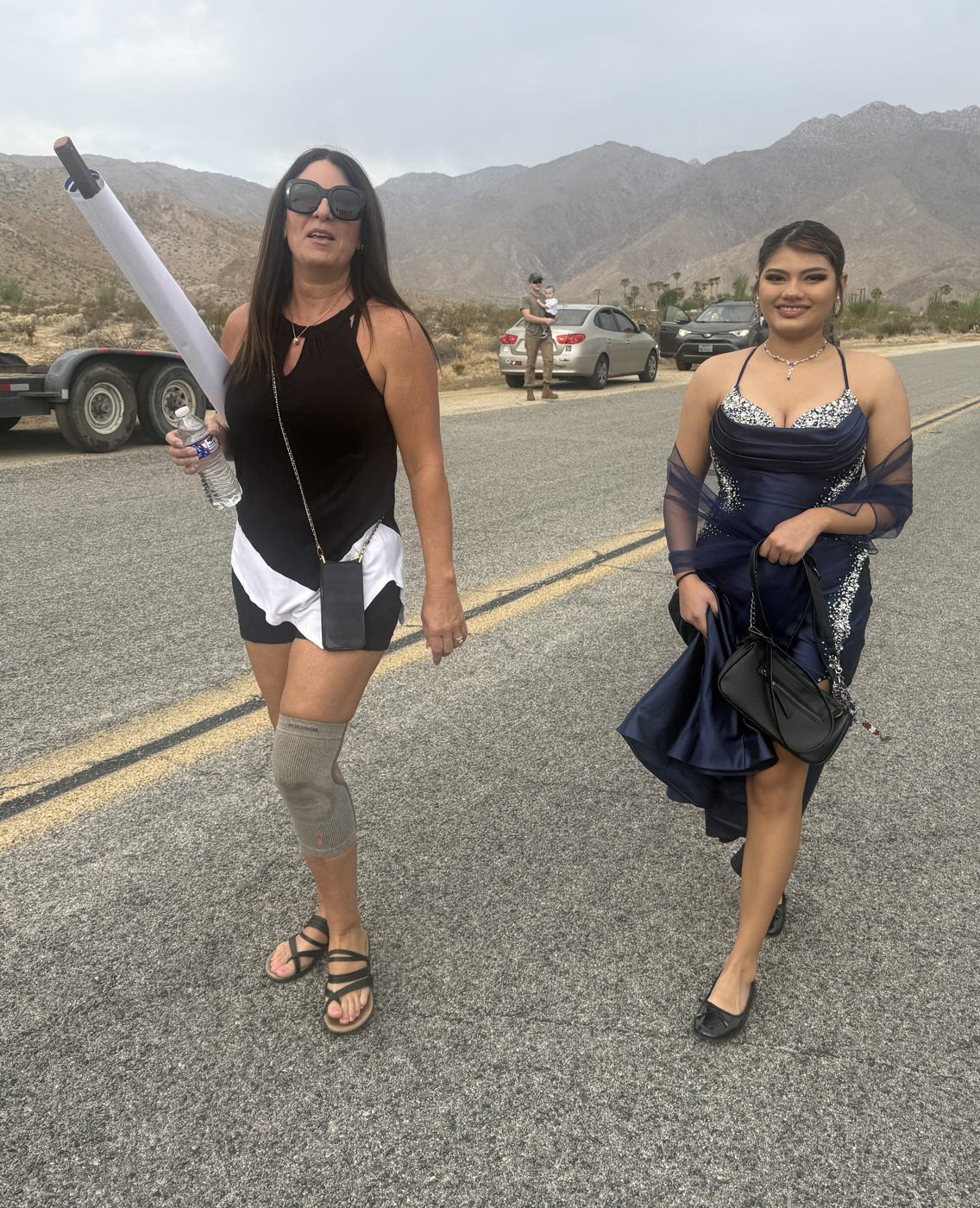 Sylvana Meeks striding up the parade lineup with a rolled banner under her arm, a Miss Borrego Springs contestant in a navy gown walking beside her